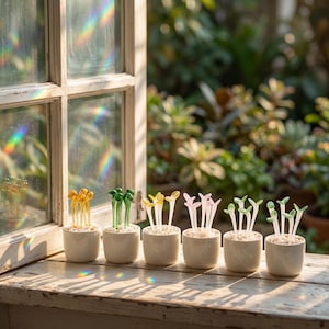 May include: Six small, white ceramic pots each hold a different colored glass plant sculpture. The glass plants are yellow, green, yellow, pink, green, and green. The pots sit on a weathered wooden surface near a window.