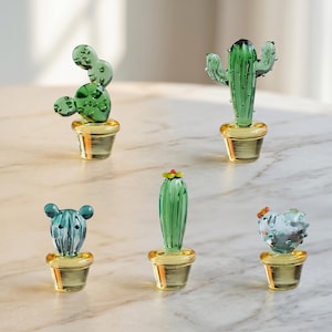 May include: Five glass cactus figurines in various shapes and shades of green, each potted in a gold-colored container. The cacti have different designs, some with spikes and others with rounded forms. The figurines are displayed on a light-colored surface.