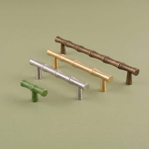 May include: Four cabinet pulls in a bamboo-inspired design, arranged in a diagonal line. The set includes one small green knob, one silver handle, one gold handle, and one dark bronze handle, each featuring segmented cylindrical detailing.