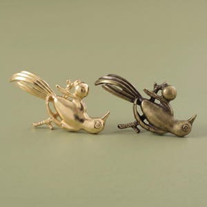 Pode incluir: Dois ornamentos de metal em forma de pássaro, um com acabamento em tom dourado polido e o outro com acabamento em tom bronze antigo. Cada pássaro é retratado pousado num galho com uma cauda longa e curva, um corpo arredondado e um bico pontiagudo.