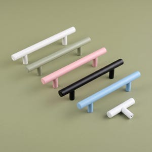 May include: A collection of six cylindrical cabinet pulls in various colors including white, sage green, soft pink, black, and light blue. Five of the pulls are long, horizontal bars with two mounting posts, and one is a small, single-post T-knob.