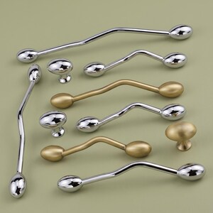 May include: A collection of cabinet hardware including several long, wavy metal pulls and small, oval-shaped knobs. The hardware features both a polished chrome finish and a matte gold finish, with each piece having rounded, bulbous ends.