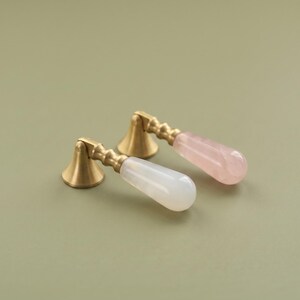 May include: Two furniture pulls featuring a brushed brass conical base and a ribbed stem. One pull has a smooth, opaque white stone handle, while the other has a translucent, pale pink rose quartz stone handle, both shaped like a teardrop.