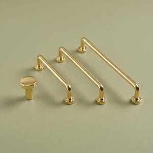 Peut inclure: Un ensemble de quincaillerie dorée composé d'un bouton de meuble rond et de trois poignées de meuble en forme de U de longueurs différentes. Chaque pièce présente une finition métallique polie et lisse avec des pieds de fixation cylindriques.