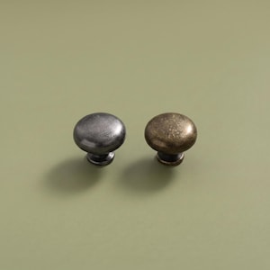 Könnte beinhalten: Zwei runde Schrankknöpfe mit glatter, gewölbter Oberfläche. Ein Knopf hat ein dunkles, gebürstetes gunmetal-graues Finish mit feinen kreisförmigen Linien, während der andere Knopf ein strukturiertes, antikes Messing-Finish mit Goldton aufweist.