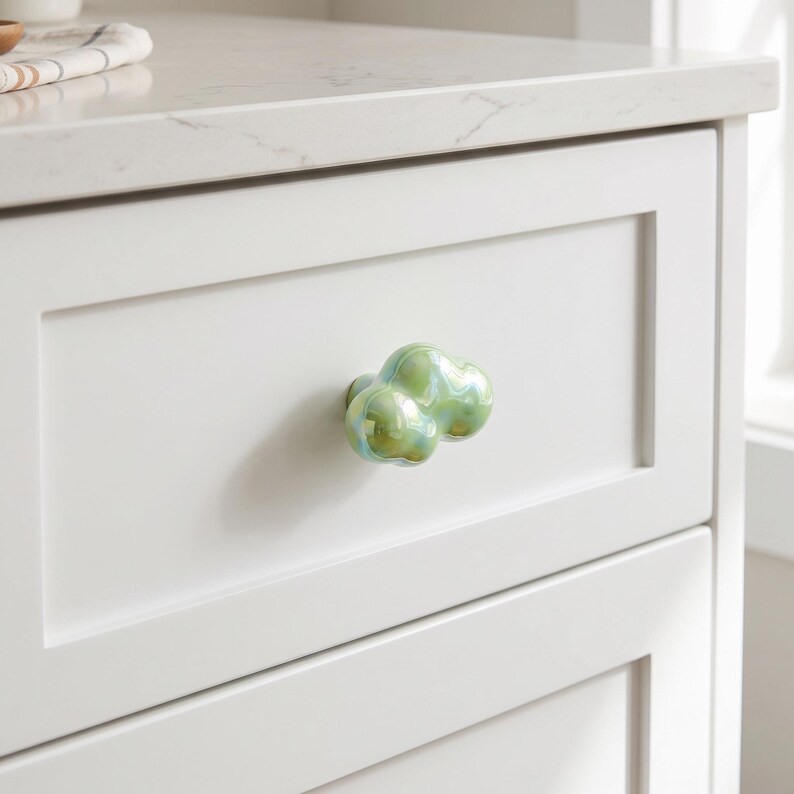 May include: A cloud-shaped cabinet knob with a glossy, iridescent finish in shades of light green and pale yellow. The knob is mounted on a white, flat-panel drawer front, featuring a smooth, reflective surface that catches the light.