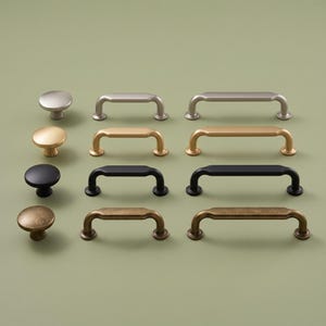 May include: A collection of twelve cabinet hardware pieces arranged in four rows. Each row features a round knob, a short handle, and a long handle in matching finishes: brushed silver, matte gold, matte black, and an antique bronze with a textured patina.