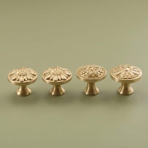 May include: Four round brass cabinet knobs arranged in a row. The two knobs on the left feature a radial petal design, while the two knobs on the right display an intricate, symmetrical embossed pattern with a beaded border around the edge.