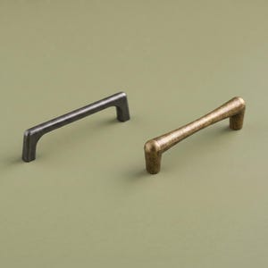 Può includere: Due maniglie per mobili in metallo sono mostrate fianco a fianco. Una maniglia presenta una finitura grigio antracite opaco scuro con un profilo rettangolare liscio. La seconda maniglia ha una finitura in ottone antico testurizzato e arrotondato.