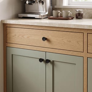 May include: A kitchen cabinet unit featuring a light oak wood grain drawer with a single matte black knob and two sage green cabinet doors below, each fitted with a matching matte black knob. The cabinet has a light-colored stone countertop.