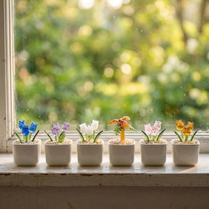 May include: Six small, square, white ceramic pots each hold a different glass flower sculpture. The flowers are blue, purple, white, orange, and yellow. The pots sit on a windowsill with a blurred green background.