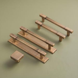 May include: A collection of antique brass hardware including one square knob and three rectangular drawer pulls. Each piece features a distinct ribbed texture on the ends and a smooth central bar, with the two larger pulls mounted on rectangular plates.