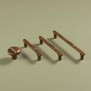 Può includere: Un set di ferramenta in ottone anticato composto da un pomello circolare e tre maniglie lineari per cassetti di varie lunghezze. Ogni pezzo presenta un profilo liscio e arrotondato con una finitura metallica scura e usurata.