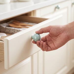 May include: A round, glossy, light blue ceramic cabinet knob attached to a cream-colored wooden drawer. The knob has a smooth, spherical shape and a reflective surface, providing a functional grip for opening the drawer in a kitchen setting.