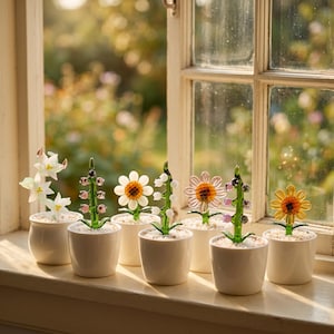 May include: A collection of glass flower sculptures in small white pots, arranged on a windowsill. The flowers include white daffodils, pink and purple beaded stems, daisies, and sunflowers. The glass flowers are set against a blurred outdoor background.