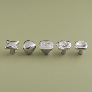 Puede incluir: Un conjunto de cinco pomos de metal plateado para armarios con formas distintas, incluyendo una estrella de cuatro puntas, un guijarro liso, un cuadrado con una espiral en el centro, un diseño floral con pétalos y un cuadrado redondeado.