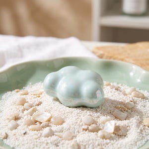 May include: A smooth, pale blue ceramic cloud-shaped object with a glossy, iridescent finish. The object is resting on a bed of small white sand and scattered light-colored pebbles inside a shallow, light green scalloped-edge bowl.