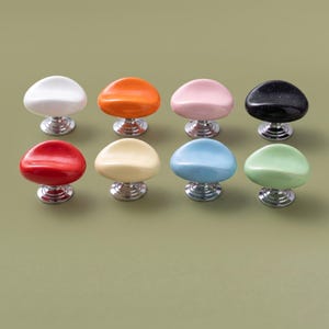Könnte beinhalten: Acht Schrankknöpfe mit glatten, abgerundeten, kieselsteinförmigen Oberteilen in Weiß, Orange, Pink, Schwarz, Rot, Creme, Hellblau und Mintgrün. Jeder Knopf verfügt über einen polierten, silberfarbenen Metallsockel mit runder Montageplatte.
