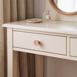 May include: A cream-colored wooden vanity table featuring a single drawer with a circular, metallic rose gold knob. The tabletop holds a small, shallow, off-white ceramic dish containing two rings and a clear glass perfume bottle with a silver cap.