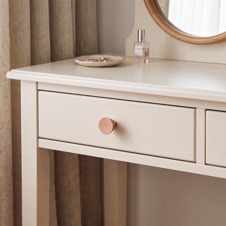 May include: A cream-colored wooden vanity table featuring a single drawer with a circular, metallic rose gold knob. The tabletop holds a small, shallow, off-white ceramic dish containing two rings and a clear glass perfume bottle with a silver cap.