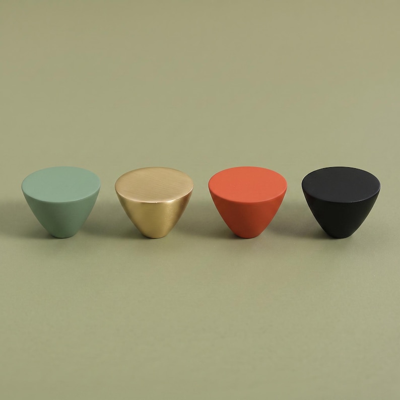 May include: Four conical cabinet knobs arranged in a row. The knobs feature a smooth, matte finish in four distinct colors: sage green, metallic gold, burnt orange, and matte black. Each knob has a flat, circular top and a tapered base.