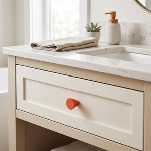 May include: A light wood bathroom vanity featuring a white marble countertop, a single drawer with a bright orange circular knob, a beige folded hand towel, a small potted succulent, and a white soap dispenser with a wooden pump mechanism.