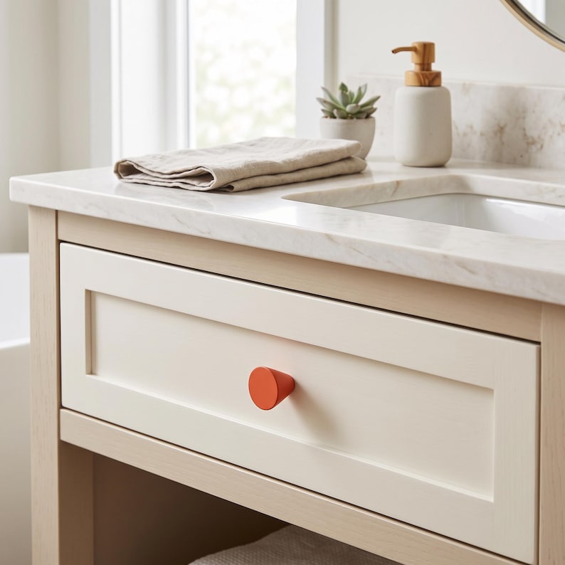 May include: A light wood bathroom vanity featuring a white marble countertop, a single drawer with a bright orange circular knob, a beige folded hand towel, a small potted succulent, and a white soap dispenser with a wooden pump mechanism.