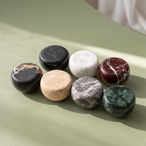 May include: A collection of seven smooth, circular stone objects in various colours and textures, including solid black, white, deep red, dark green, grey marble, beige, and a black stone with tan veining, all arranged in a cluster.