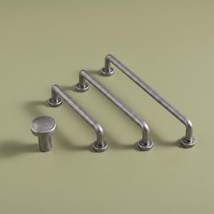 Puede incluir: Un juego de herrajes para muebles que consta de un pomo redondo y tres tiradores de barra alargados. Cada pieza presenta un acabado metálico oscuro y desgastado con un diseño cilíndrico liso y bases de montaje circulares para fijar.