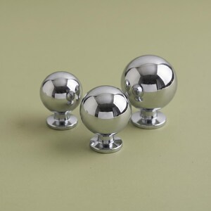 May include: Three spherical cabinet knobs with a polished chrome finish. Each knob features a smooth, reflective, ball-shaped head mounted on a circular base. The set includes one large knob and two smaller knobs, all displaying a metallic sheen.