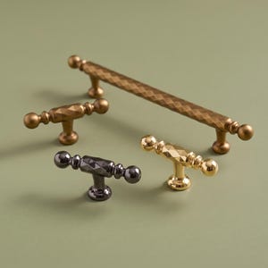 May include: A collection of four metal cabinet hardware pieces including one long bar pull and three T-shaped knobs. The hardware features a textured diamond-patterned center with smooth spherical ends and a central mounting post in brass and black.