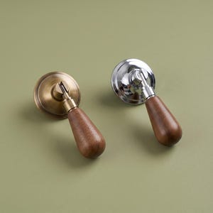 May include: Two teardrop-shaped wooden cabinet pulls with metal bases. One pull features a brushed brass circular backplate, while the other has a polished chrome circular backplate. Both handles have a smooth, brown wood finish.