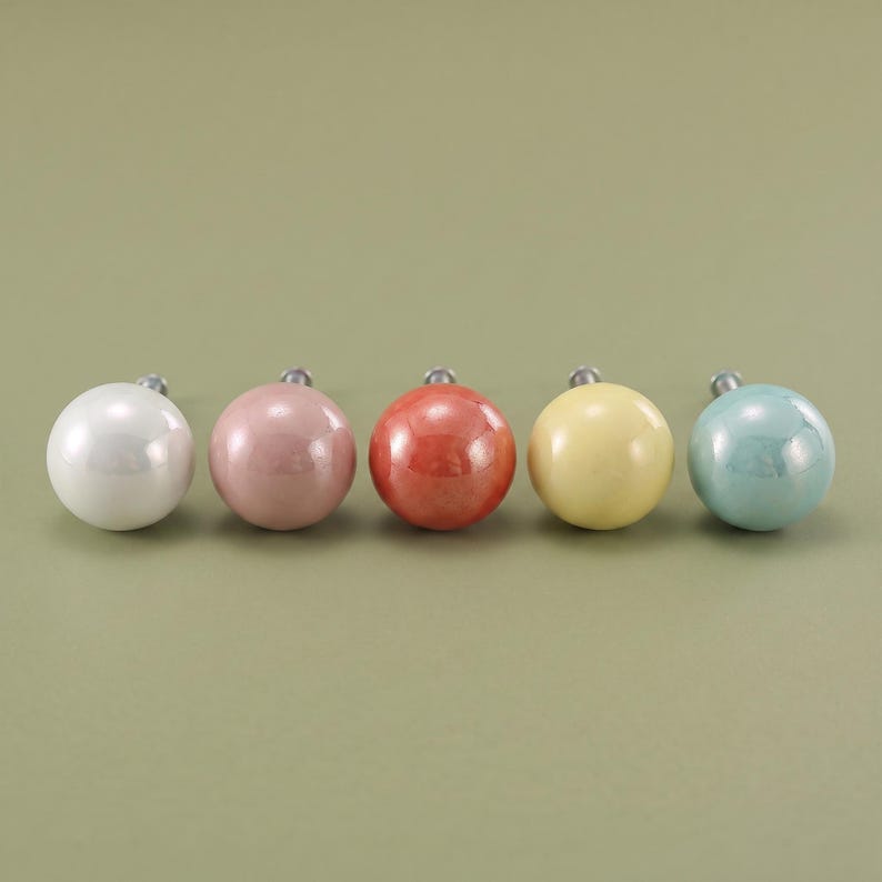 May include: A set of five spherical cabinet knobs arranged in a horizontal row. The knobs feature a smooth, glossy finish in distinct colors: white, dusty pink, coral red, pale yellow, and light teal, each with a small metal mounting screw.