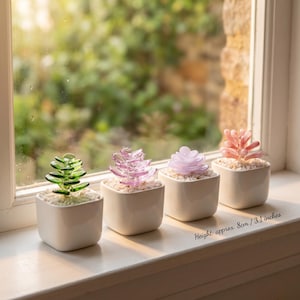 May include: Four small, square white ceramic pots each hold a different colored glass succulent. The succulents are green, pink, lavender, and coral. The height is approx. 3.1 inches. The pots sit on a white windowsill with a blurred green background.