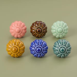 Puede incluir: Un conjunto de seis pomos de cerámica redondos dispuestos en dos filas. Cada pomo presenta un borde festoneado en forma de flor y un centro circular elevado. Los colores incluyen rosa pastel, marrón oscuro, verde menta claro, amarillo mostaza, azul real y verde azulado.