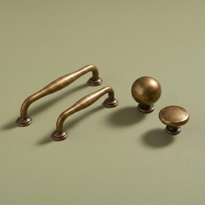 May include: Two antique brass cabinet pulls with a curved, tapered design and two matching round cabinet knobs. The hardware features a weathered, dark metallic finish with subtle highlights, showcasing a classic shape for furniture or cabinetry.