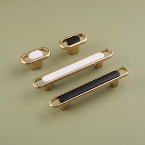 May include: A collection of four cabinet pulls featuring a metallic gold finish. Two pulls are small oval knobs with white or black inserts. Two pulls are long, slender handles with rounded ends, one with a white insert and one with a black insert.