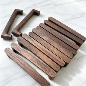 May include: A collection of dark brown wooden cabinet handles with a smooth, elongated, and curved design. The set includes several horizontal handles arranged in a row and two upright handles positioned separately to the left side.
