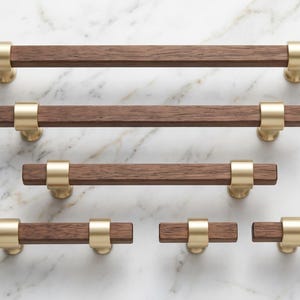 May include: A collection of five cabinet pulls in varying lengths, each featuring a dark brown rectangular wooden bar accented by two brushed gold metal mounting brackets. The hardware is arranged in a horizontal layout with different sizes.