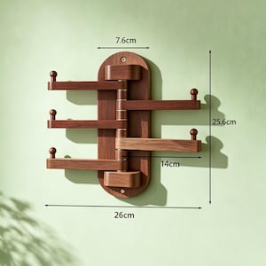May include: A wooden wall-mounted coat rack with a dark brown finish. The rack is 26 cm wide and 25.6 cm tall, featuring three pivoting arms with rounded knobs. The top section is 7.6 cm wide. The rack is designed for hanging items.