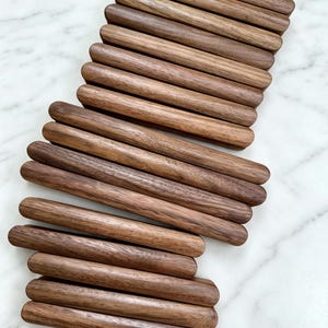 May include: A collection of fifteen cylindrical wooden dowels arranged in a staggered, overlapping pattern. Each dowel features a smooth, polished finish with visible natural wood grain patterns in varying shades of medium to dark brown.