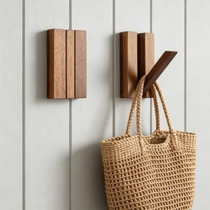 Puede incluir: Dos ganchos rectangulares de pared hechos de madera marrón, cada uno con tres listones verticales. Un gancho está vacío, mientras que el otro sostiene un bolso de paja tejido en color beige con asas trenzadas colgado de su brazo de madera inclinado.