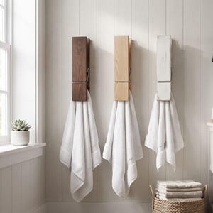 May include: Three wall-mounted towel hooks shaped like oversized wooden clothespins, each holding a white towel. The hooks are finished in dark brown, natural light wood, and a distressed white paint, featuring a silver metal spring detail.