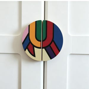 May include: A pair of semi-circular cabinet pulls mounted on white doors. Each half features a bold, geometric pattern with segments of pink, orange, green, red, blue, purple, and cream, all outlined in thick black lines to form a cohesive design.