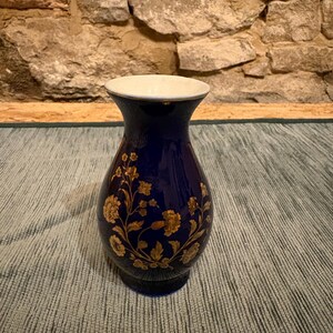 Könnte beinhalten: Eine dunkelblaue Keramikvase mit weißem Rand und goldenem Blumendesign. Die Vase hat einen abgerundeten Körper und einen schmalen Hals. Das goldene Blumenmuster ist detailliert und bedeckt einen großen Teil der Oberfläche der Vase.