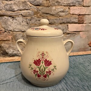 Könnte beinhalten: Cremefarbener Keramiktopf mit Deckel und zwei Griffen. Der Topf hat ein Blumendesign mit roten Tulpen und grünen Blättern. Der Deckel hat einen kleinen Knopf und ein passendes Blumenmuster. Der Topf steht auf einer blauen Oberfläche.