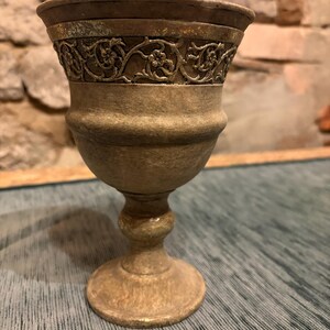 Könnte beinhalten: Ein dekorativer Kelch mit detailliertem floralem Design am Rand. Der Kelch hat eine gedämpfte Goldfarbe mit strukturierter Oberfläche. Sockel und Stiel sind ebenfalls goldfarben und erzeugen eine Vintage-Ästhetik.