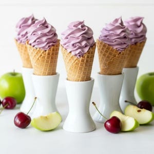 May include: Six ice cream cones filled with lavender-colored soft serve, presented in white holders. Green apple slices and cherries are scattered around the cones. The cones have a waffle texture.