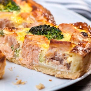 Puede incluir: Una porción de quiche con salmón y brócoli sobre un plato blanco. La quiche tiene una corteza dorada y un relleno cremoso amarillo. Se ven trozos de salmón y floretes de brócoli.