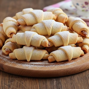 Puede incluir: Una tabla de madera llena de pasteles frescos en forma de media luna. Las galletas doradas están espolvoreadas con azúcar glas, creando un dulce y apetitoso manjar. Perfecto para un postre o un tentempié.